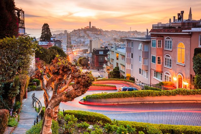 Curvy road winds downhill, surrounded by colorful houses and lush greenery, under a vibrant sunset sky in a bustling cityscape.