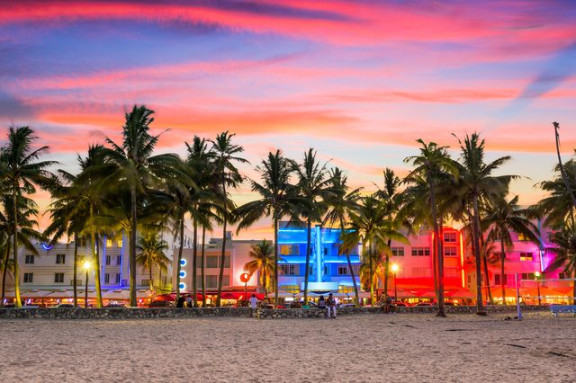 Palm trees stand silhouetted against colorful sunset skies, framing vibrant neon-lit buildings, with a sandy beach in the foreground.