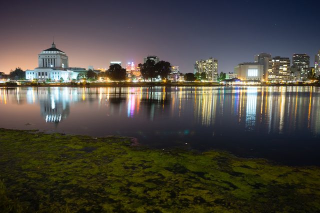 Buildings reflect in a calm lake under a night sky, with colorful city lights creating shimmering patterns on the water's surface.