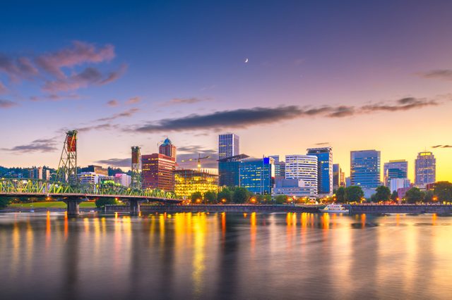 City skyline reflects on a calm river at sunset, with a bridge spanning across and colorful buildings illuminated against a transitioning evening sky.