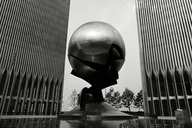 A large metallic sphere sits statically between two tall buildings, surrounded by trees and reflected in the pool below.