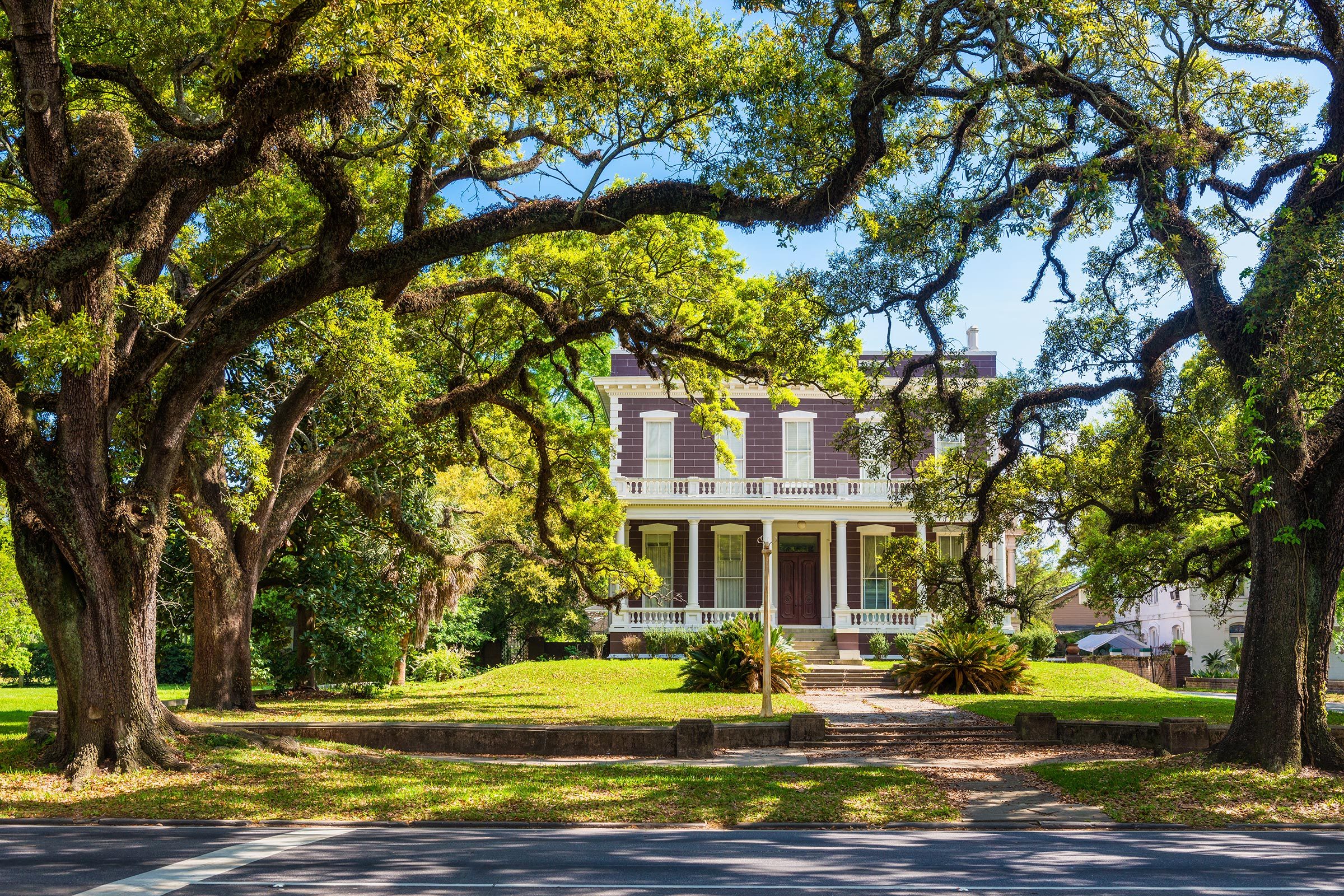 A grand two-story house stands amidst large, arching oak trees and lush greenery in a sunny, park-like setting.