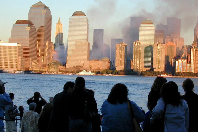 Spectators observe smoke rising behind skyscrapers across a river during sunset, creating a dramatic urban scene filled with anticipation and tension.