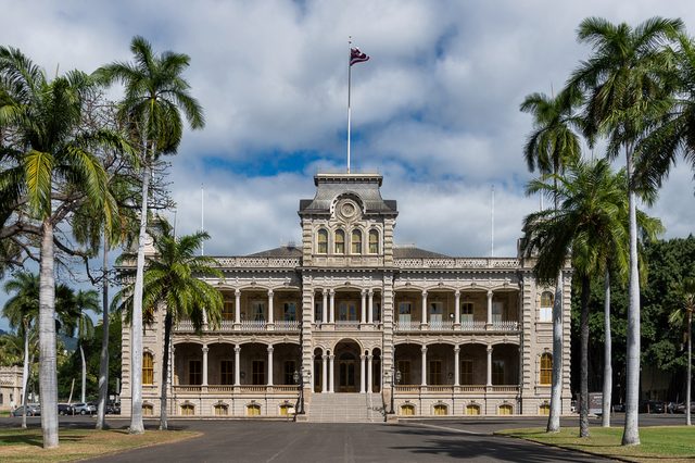 A historic palace stands majestically, flanked by tall palm trees, under a partly cloudy sky with a flag flying atop.