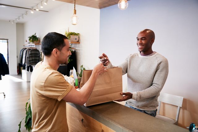 Two people exchange a shopping bag over a counter in a clothing store, smiling warmly, with racks and plants in the background.