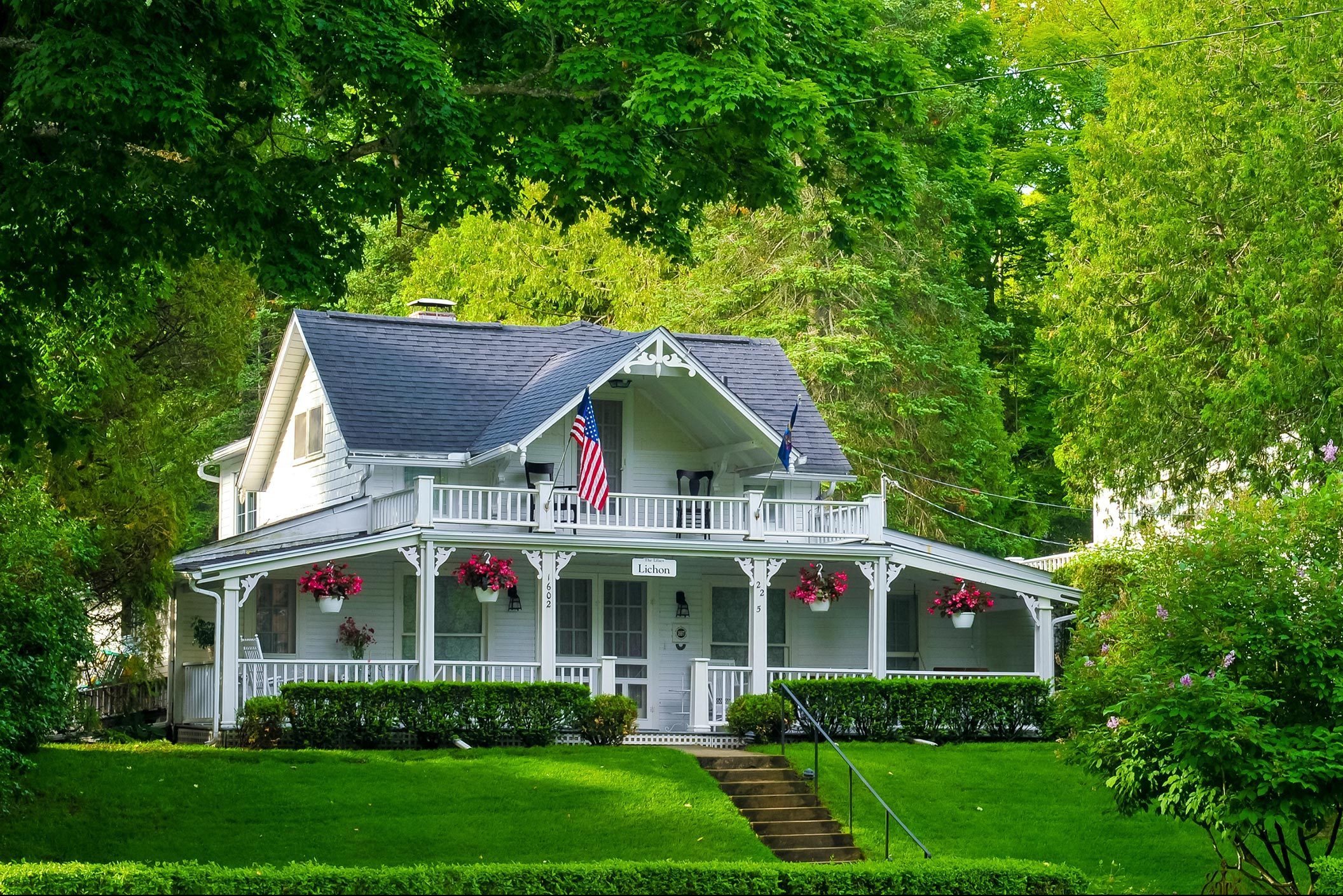 White house with American flag, surrounded by lush greenery, flowers hanging from porch, brick path leading through well-maintained lawn in a wooded setting.