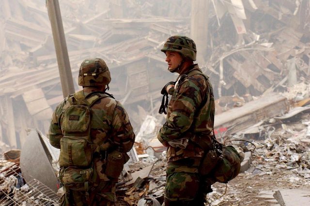 Soldiers stand amid debris, surveying destruction. The scene is chaotic, with scattered ruins and dust creating a somber atmosphere.