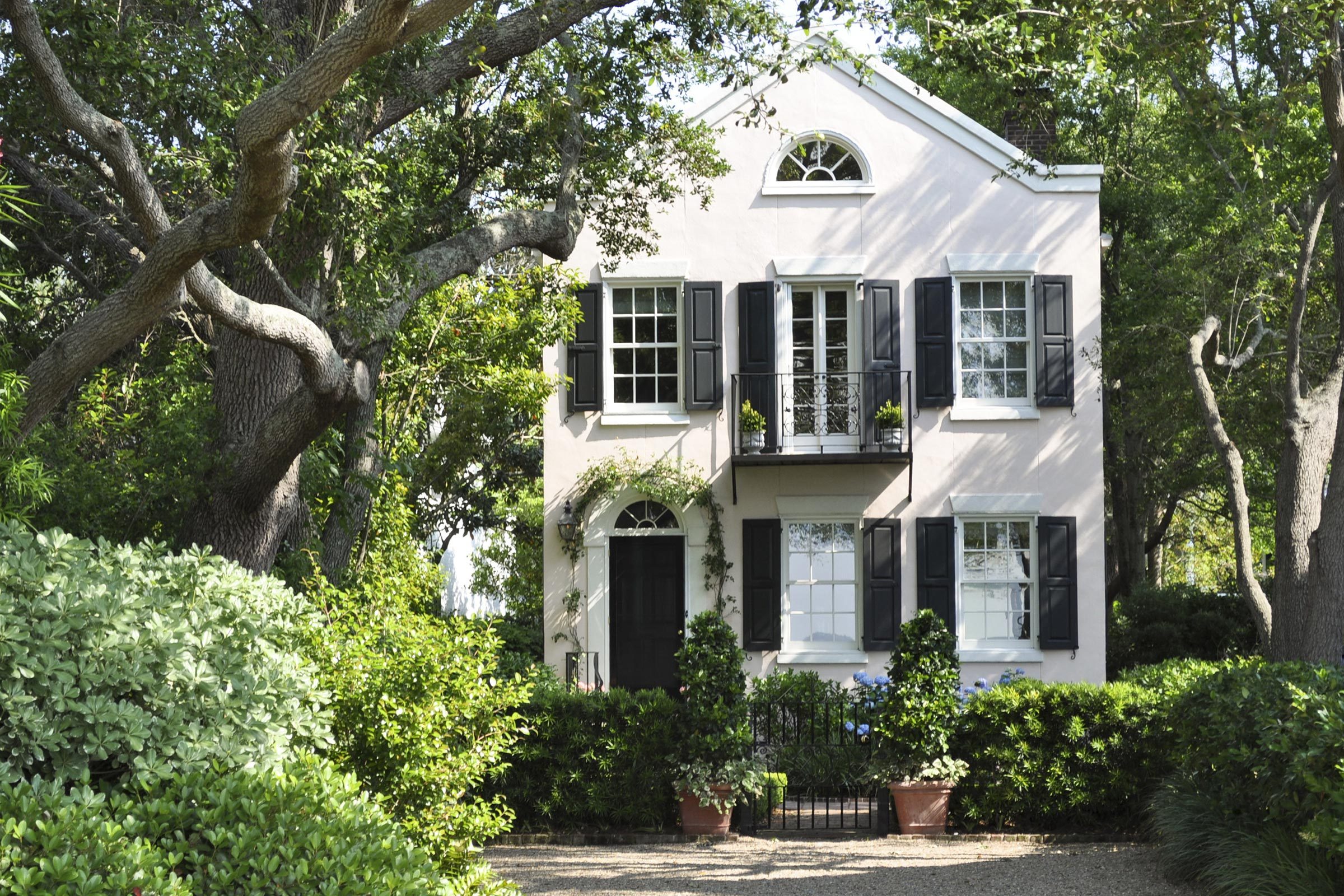 A two-story house sits among lush trees and shrubs, featuring white walls, black shutters, and a small balcony with potted plants.