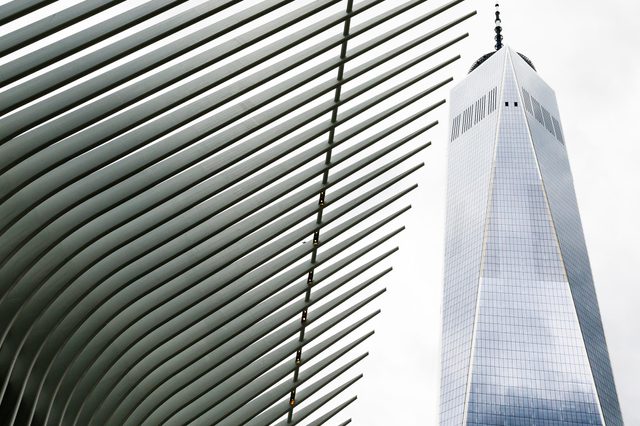 A tall skyscraper stands beside ribbed architectural beams, both reaching upwards against a cloudy sky.