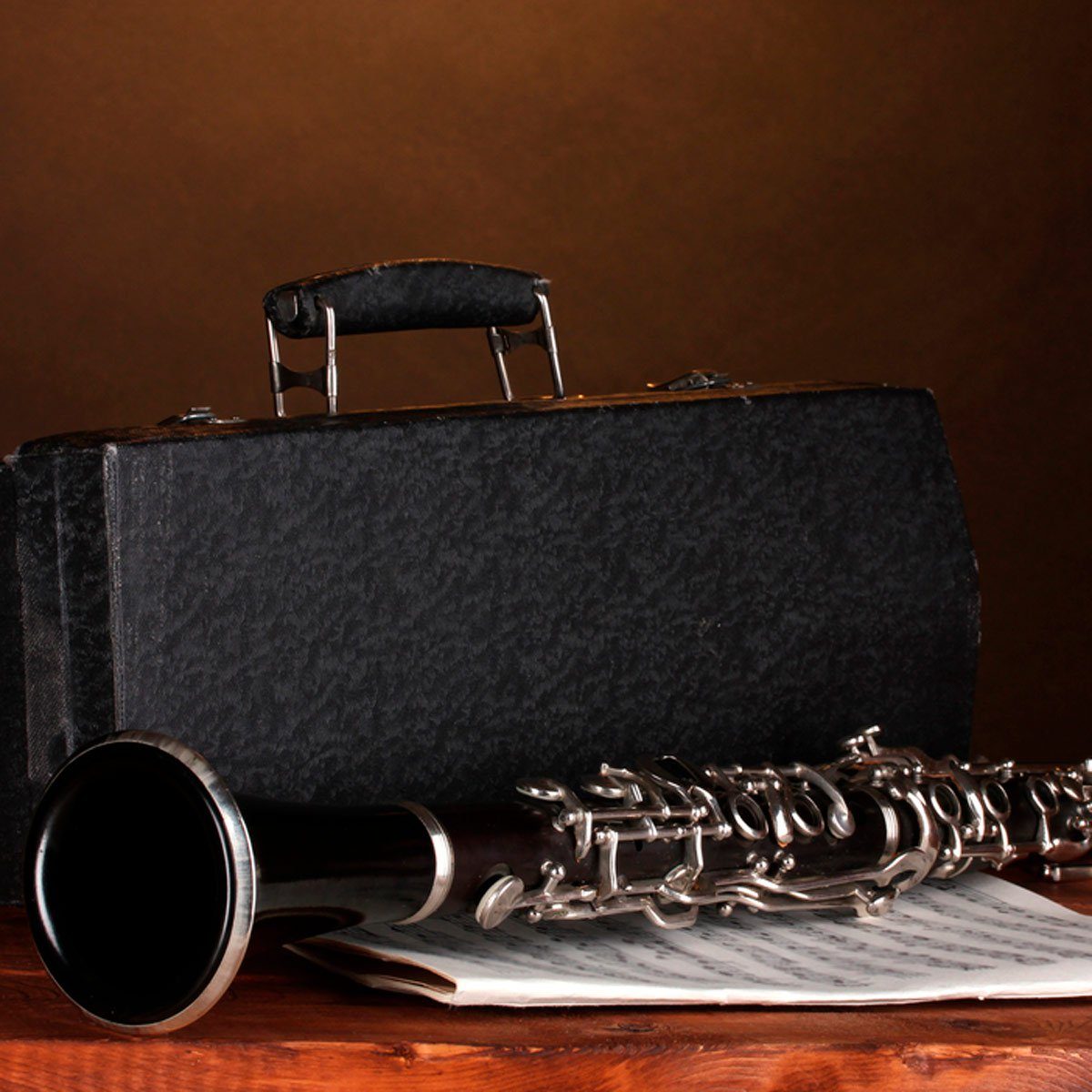 Clarinet rests on sheet music beside a black case, all placed on a wooden surface in a dimly lit room.