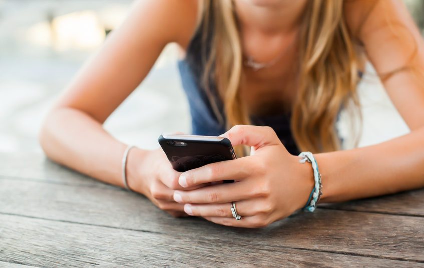 Hands hold a smartphone, resting on a wooden surface, outdoors. A bracelet and ring are visible on the person's hand and wrist.