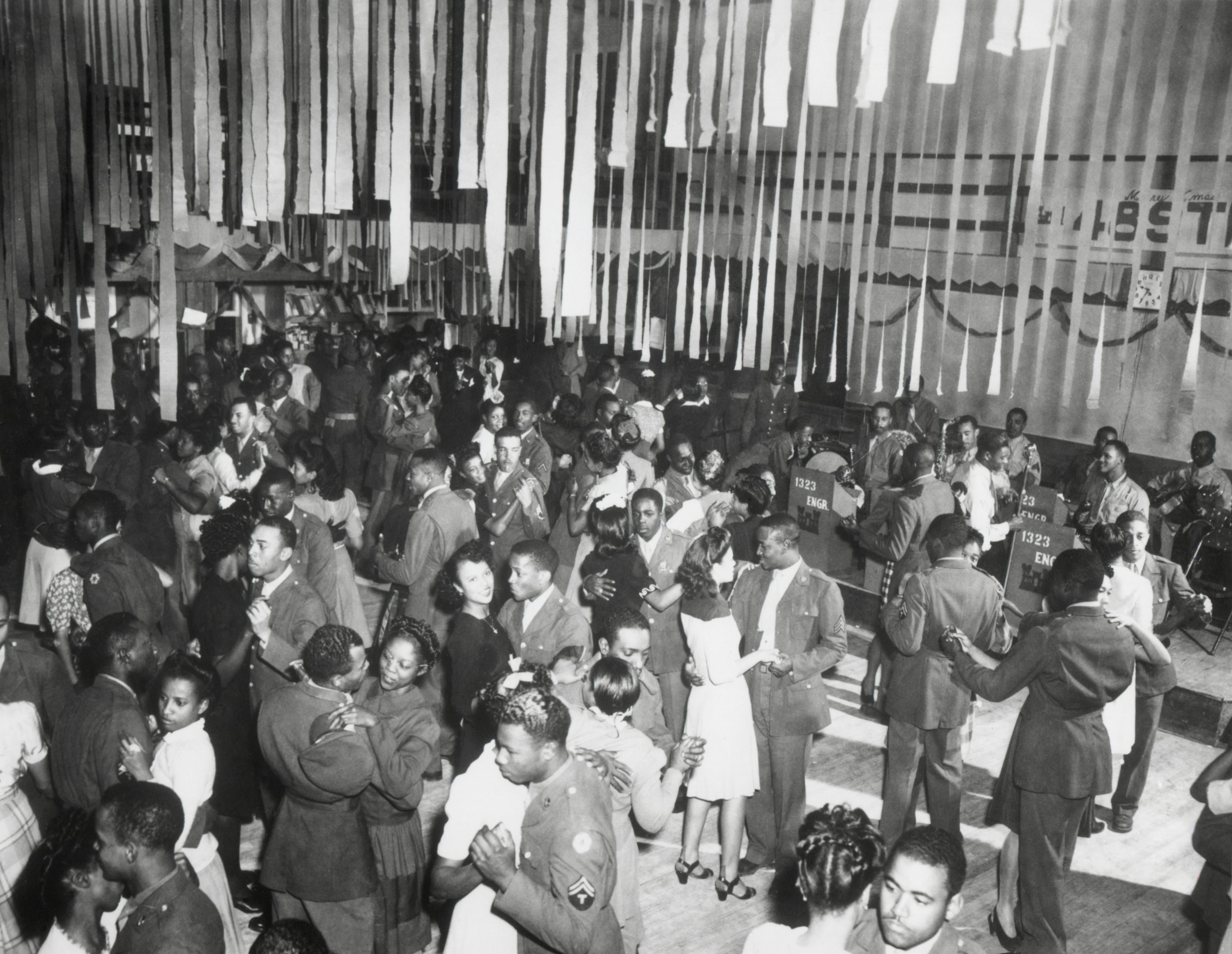 Couples dance closely on a crowded floor; military uniforms and formal attire visible under hanging streamers, with a live band playing in the background.