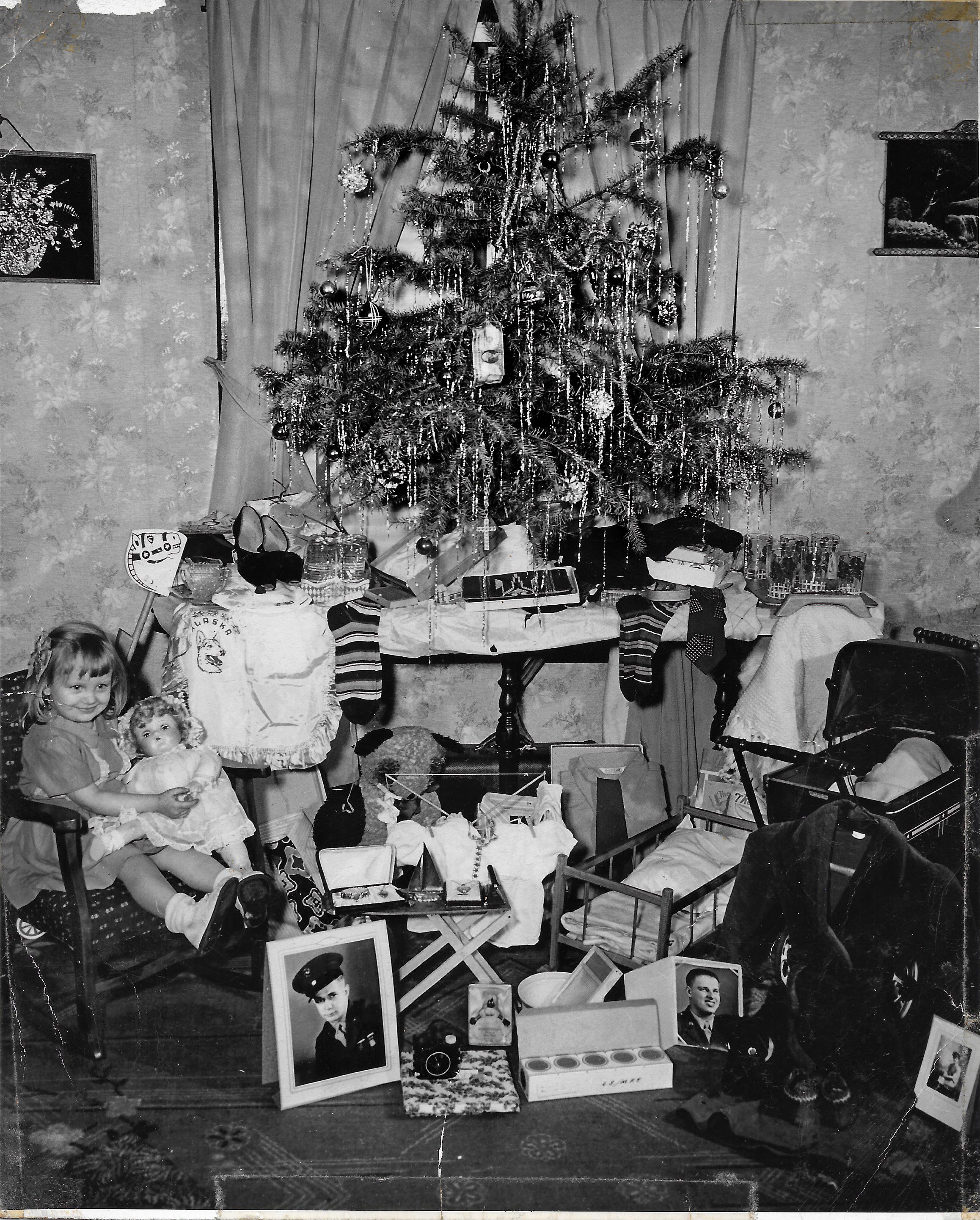 Girl holds doll, sitting beneath a decorated Christmas tree surrounded by gifts and framed photographs in a cozy, curtain-draped room.
