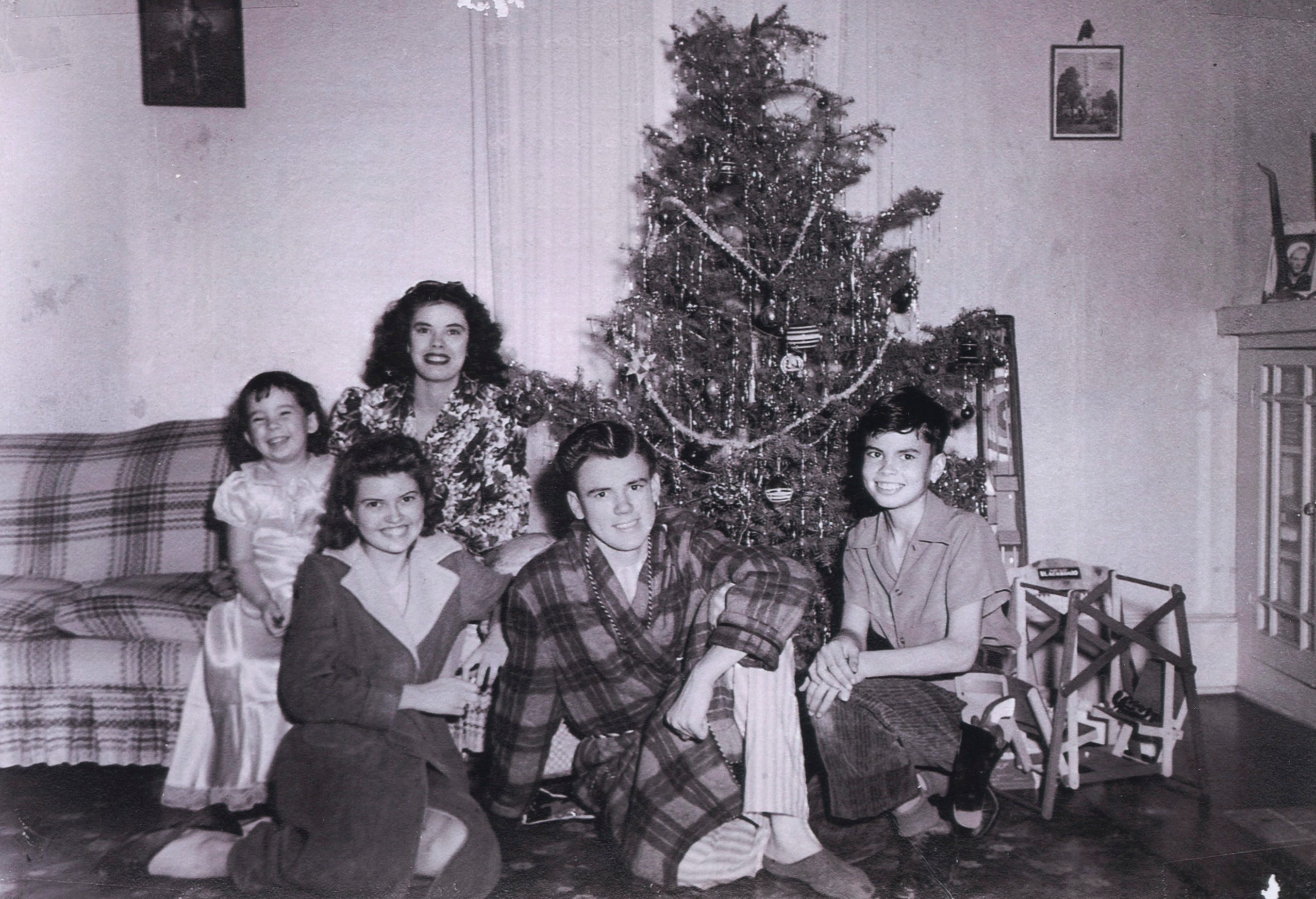 A family sits smiling by a decorated Christmas tree in a living room with a plaid couch and framed pictures on the walls.