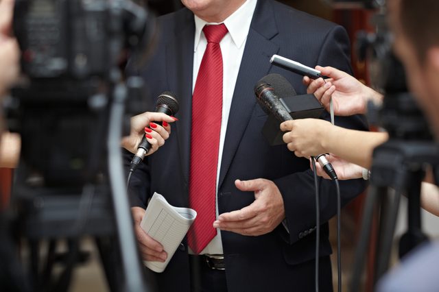 Person in a suit speaks to journalists holding microphones, surrounded by cameras and reporters in a press conference setting.