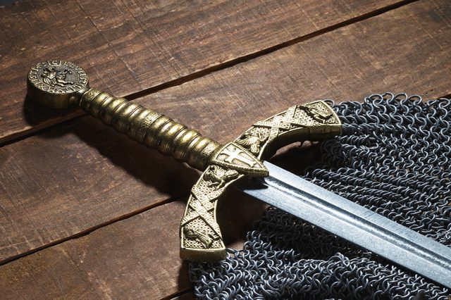 A decorative sword with a gold hilt rests on chainmail atop a wooden surface, showcasing intricate medieval designs.