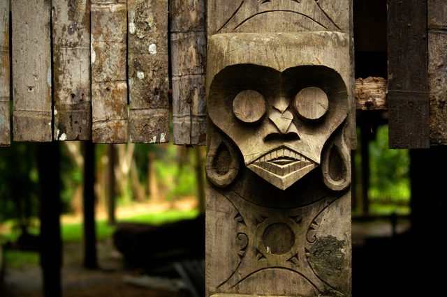 Wooden mask carved into a post, featuring circular eyes and geometric designs, set against a lush forest background.