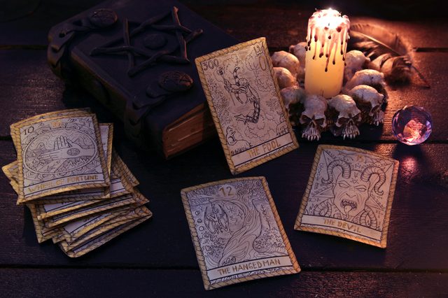 Tarot cards lie scattered near an ornate book and lit candle surrounded by skulls, creating a mystical atmosphere on a dark wooden table.
