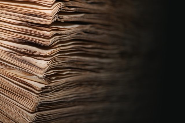 A stack of aged, yellowed papers sits in dim light, edges curled and layered, creating a textured pattern against a dark background.