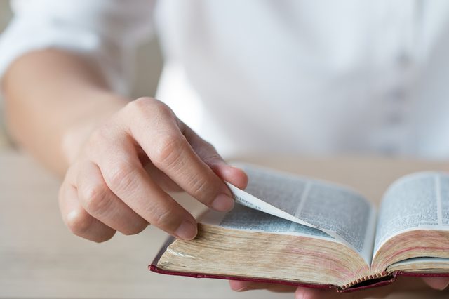 Hand turns a page in an open book, resting on a wooden table in a brightly lit room.