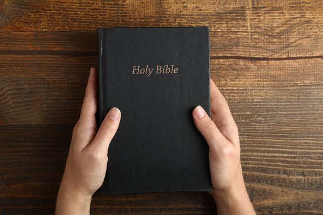 Hands hold a black book titled "Holy Bible" on a wooden table.