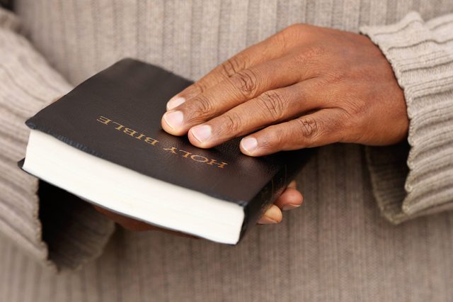 Hands hold a closed black book labeled "Holy Bible," against a gray, textured sweater background.