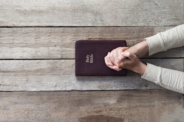 Hands clasped in prayer over a closed Bible on a rustic wooden table.