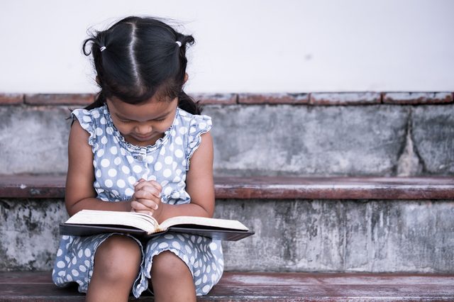 Child reads intently, sitting on stone steps, wearing a blue polka dot dress with hands clasped over a book.