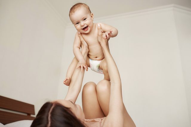 A baby, smiling and airborne, is held playfully by an adult in a minimalist room setting.