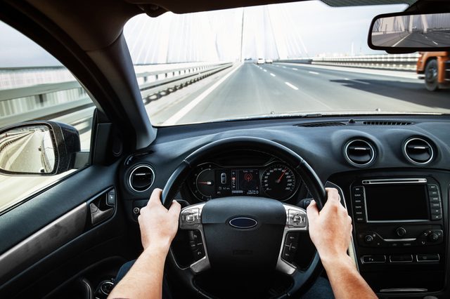 Hands grip a steering wheel, driving on a bridge with cables and lanes ahead, viewed from inside a car.
