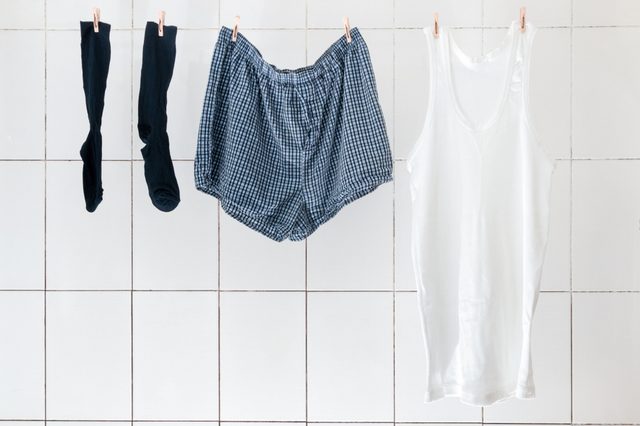 Clothes hang drying on a line, including black socks, checkered shorts, and a white tank top against a tiled wall background.