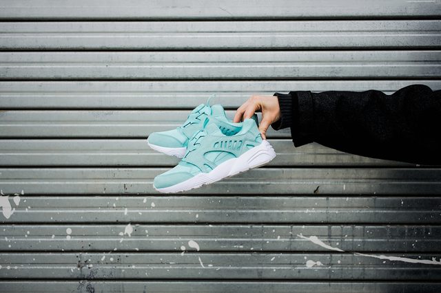 Hand holds turquoise sneakers against a metal, horizontally-striped wall with white paint splatters.