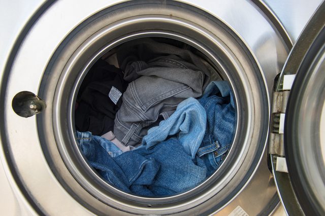 Clothes fill a stainless steel washing machine drum, displaying various shades of denim and dark fabric, set in a laundry room environment.