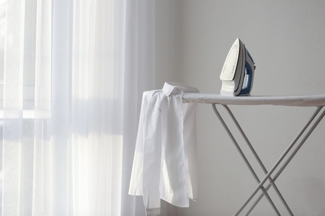 Iron rests on ironing board with a white shirt, near a sunlit window in a minimalist room.