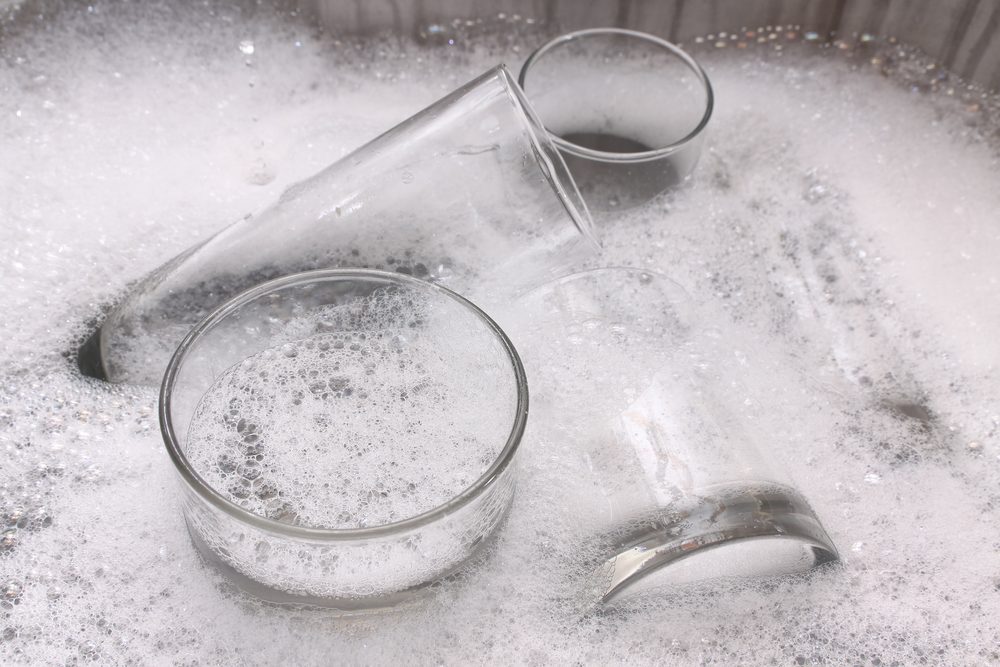 Glasses soak in soapy water, surrounded by foam, in a kitchen sink.