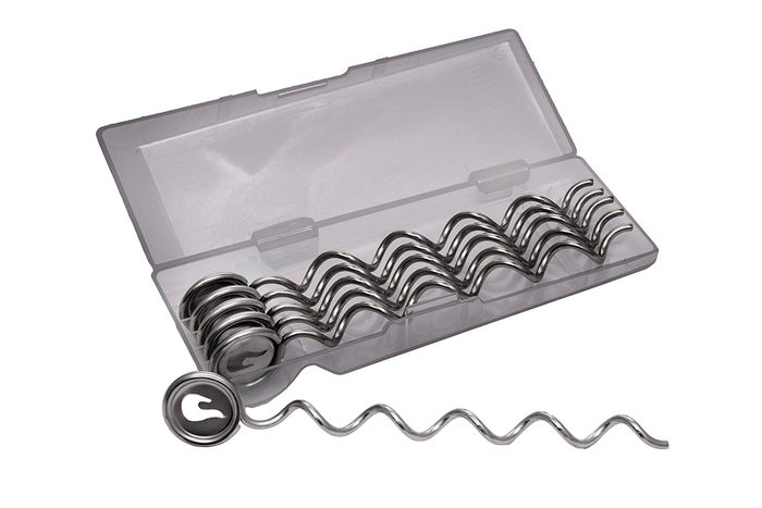 Metal spiral garden stakes rest in a clear plastic box; one stake is partially outside. The setting appears to be a product display.