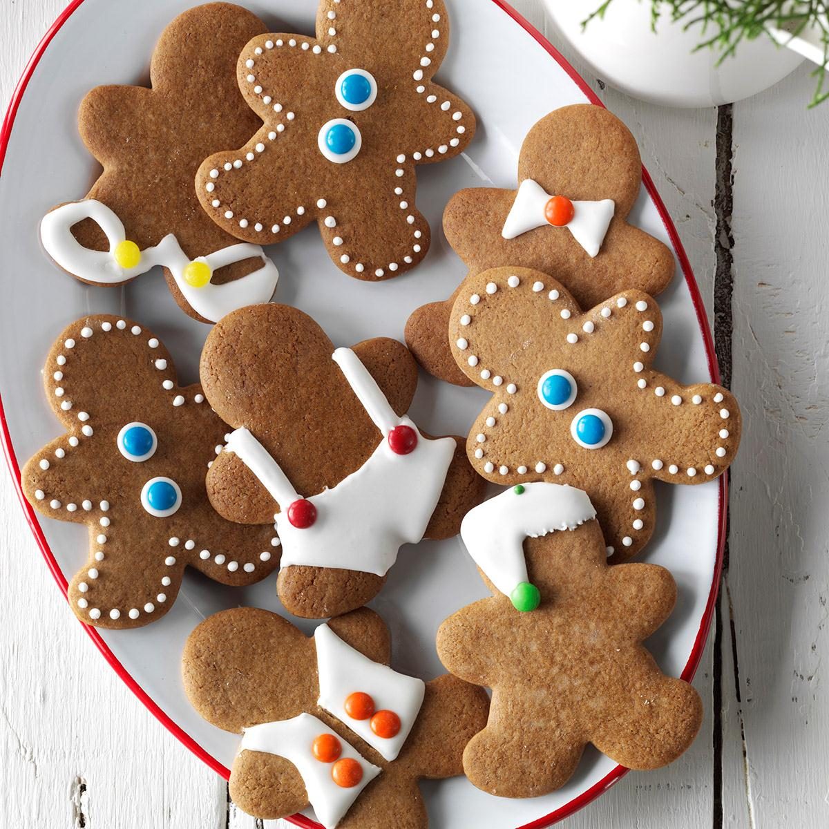Gingerbread cookies decorated with icing and candy buttons lie on a red-rimmed plate atop a white wooden table.