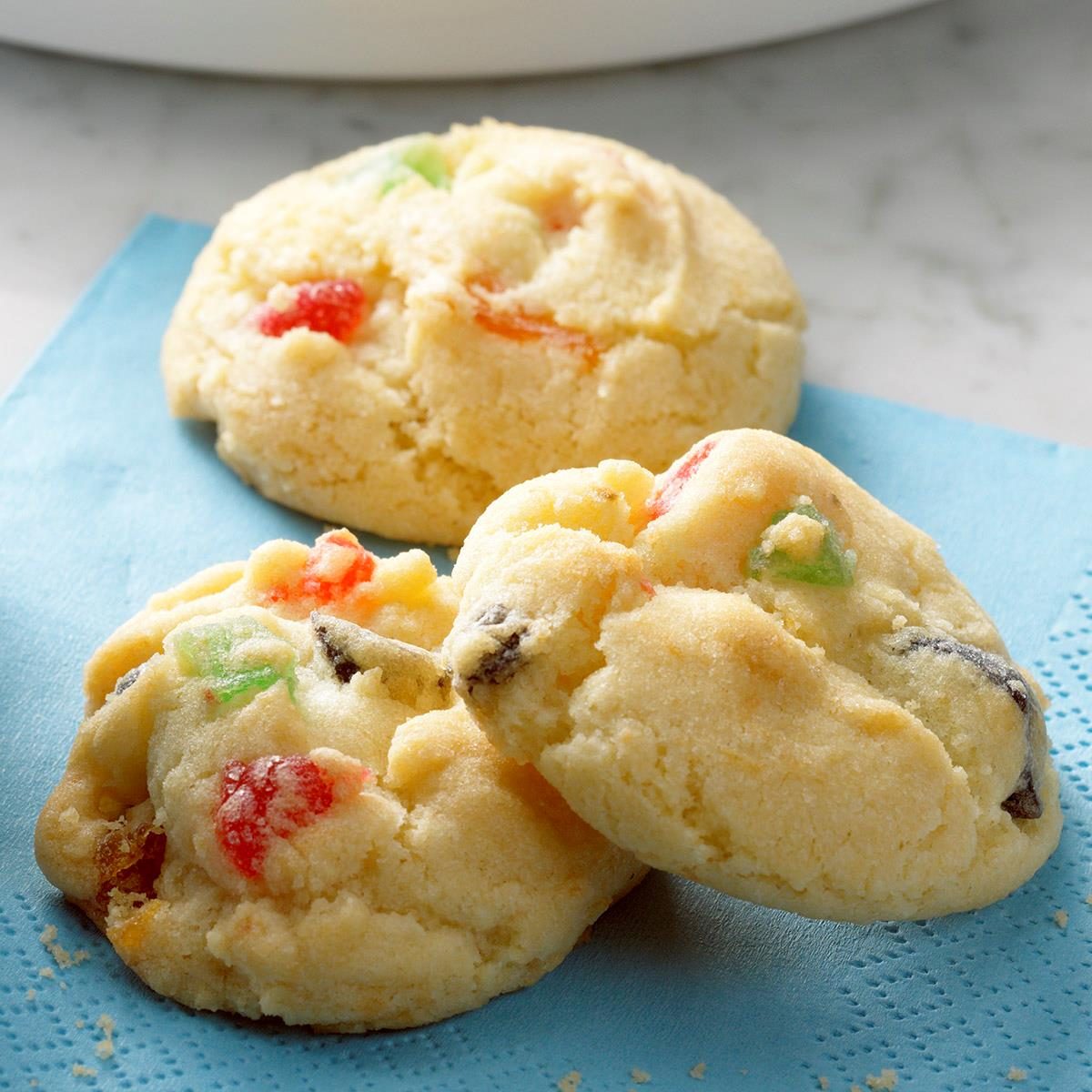Cookies sit on a blue napkin, showing colorful candied fruit pieces, with a marble surface in the background.