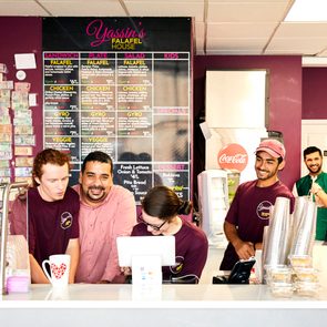 Employees smile behind the counter, interacting with customers in a falafel restaurant. A menu board listed with options on the wall adds context.