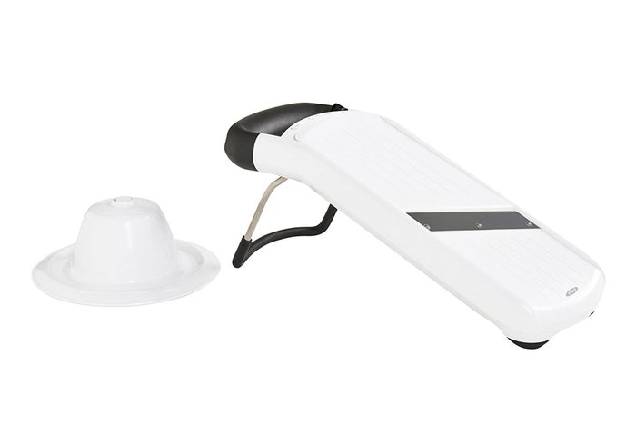 A white mandoline slicer rests on a stand, with a food holder nearby, on a plain white background.