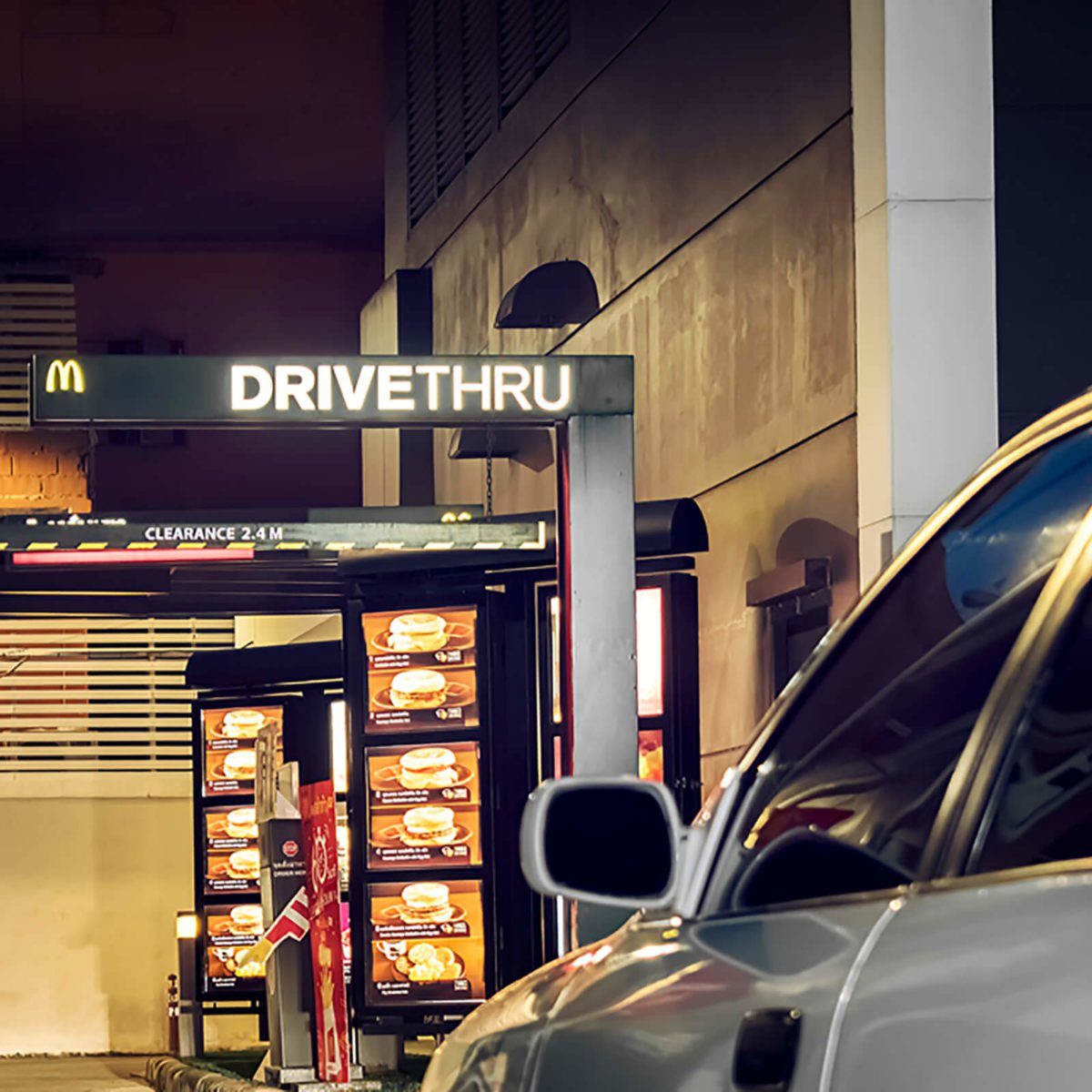 Car waiting at a drive-thru, illuminated by restaurant menu boards, under signage reading "DRIVETHRU" with clearance marked at 2.4 meters.