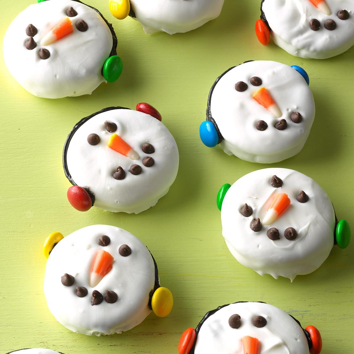 Cookies decorated as snowman faces with candy eyes and noses, lie on a pastel green surface.