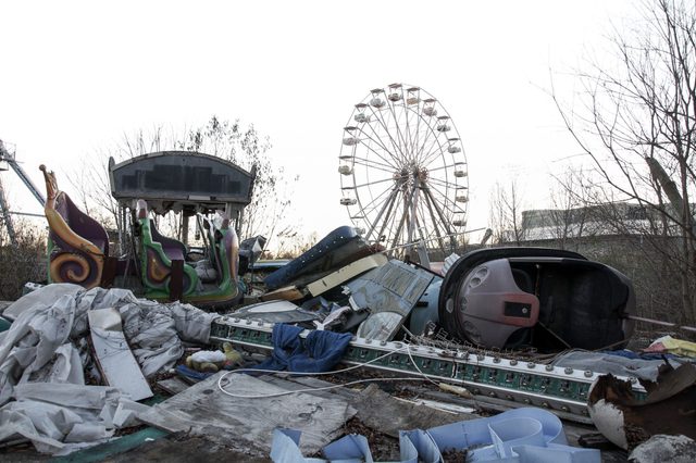 Abandoned amusement park rides lie scattered and deserted amidst overgrown trees with a Ferris wheel in the background.