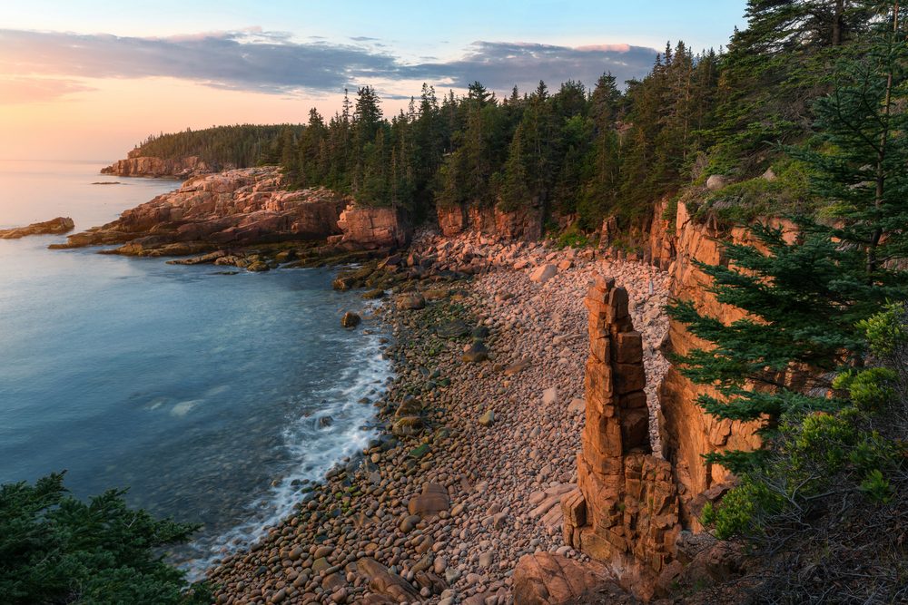 Rugged cliffs rise above a rocky shoreline, bordered by dense evergreen forests under a colorful sunset sky.