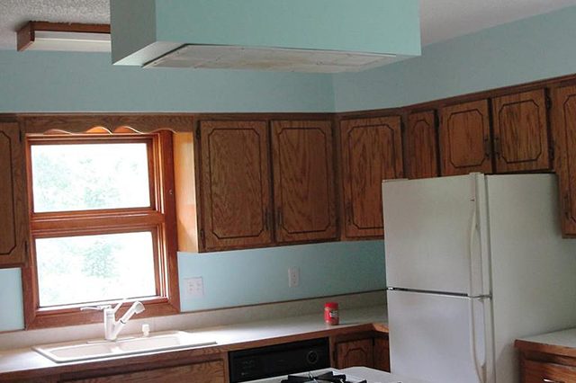 Stove sits on a kitchen island, surrounded by wooden cabinets, near a window and refrigerator in a softly lit room.