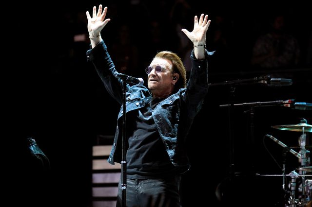 A man stands on stage, arms raised, wearing sunglasses and a denim jacket, in front of a microphone and musical equipment.