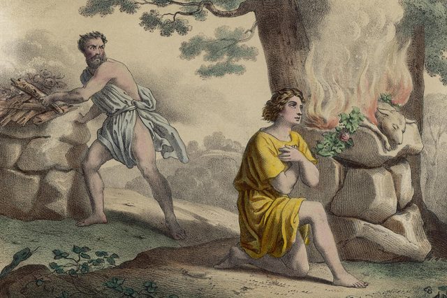 Two men perform a ritual; one kneels in prayer, the other prepares a burning altar with a lamb in a wooded landscape.