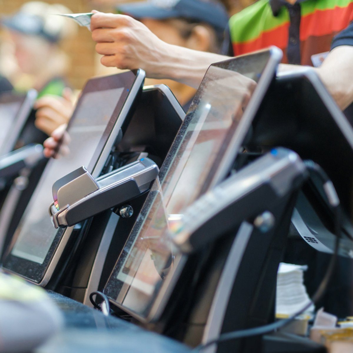 Touchscreen registers processing transactions; a hand inserts a card. The setting is a busy retail or restaurant checkout area.