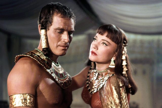 Two people adorned in elaborate ancient Egyptian attire stand close, gazing at each other, in a richly decorated room with draped fabric.