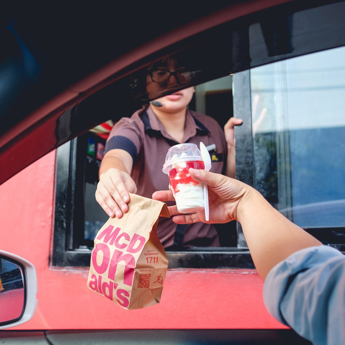 Person hands McDonald's bag and sundae to driver through a red drive-thru window.